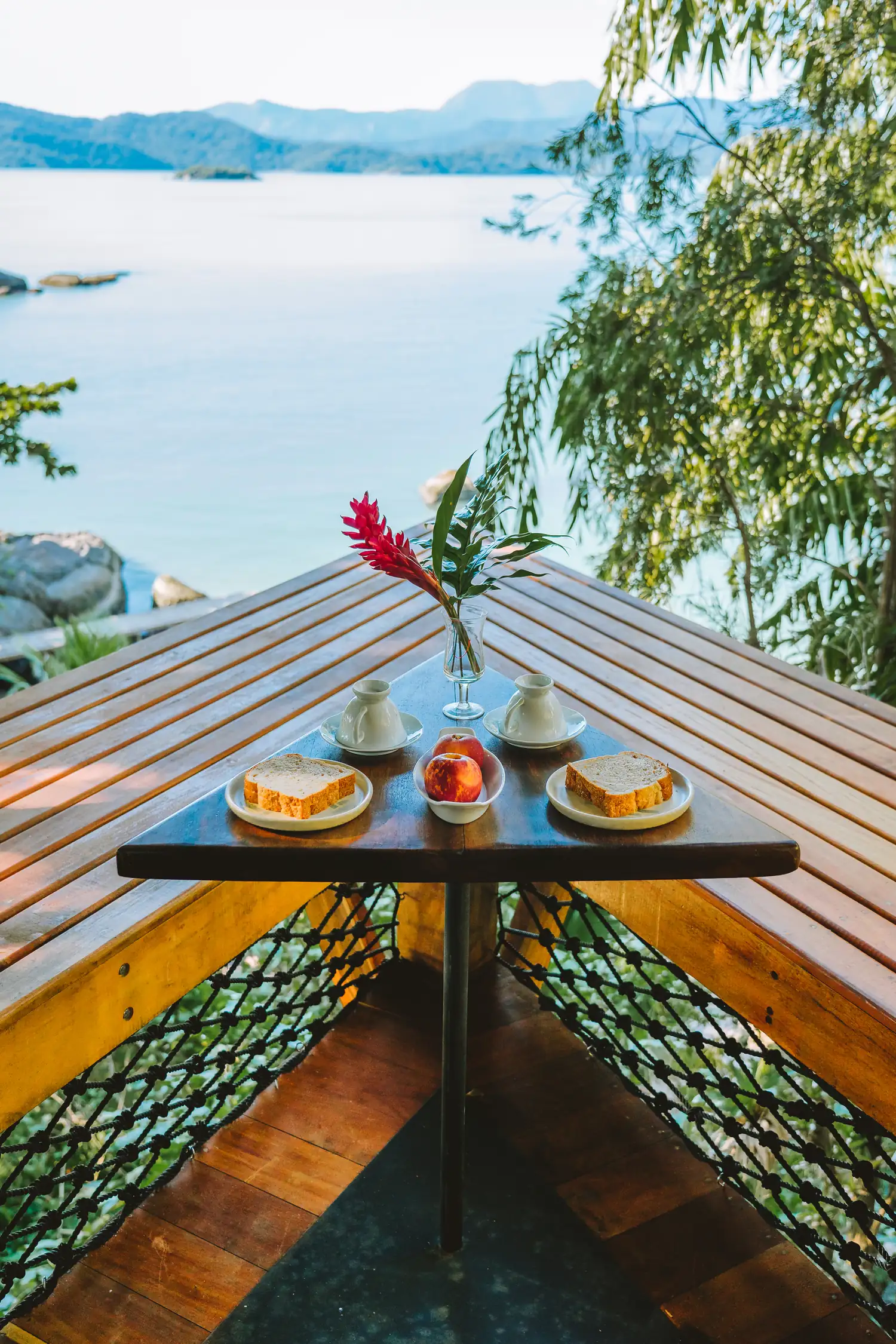 Café da manhã servido na mesa de madeira da proa, com pães, frutas e a baía clara ao fundo